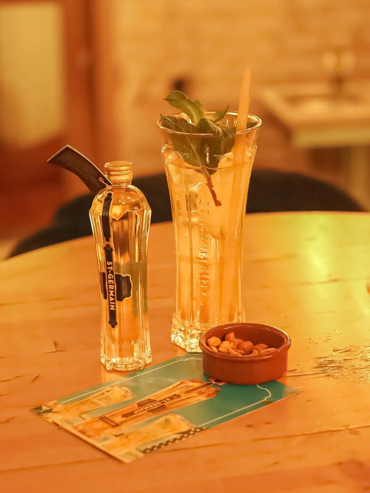 Cocktail St-Germain à la menthe dans un grand verre ciselé, mignonnette de liqueur et coupelle de cacahuètes sur une table en bois