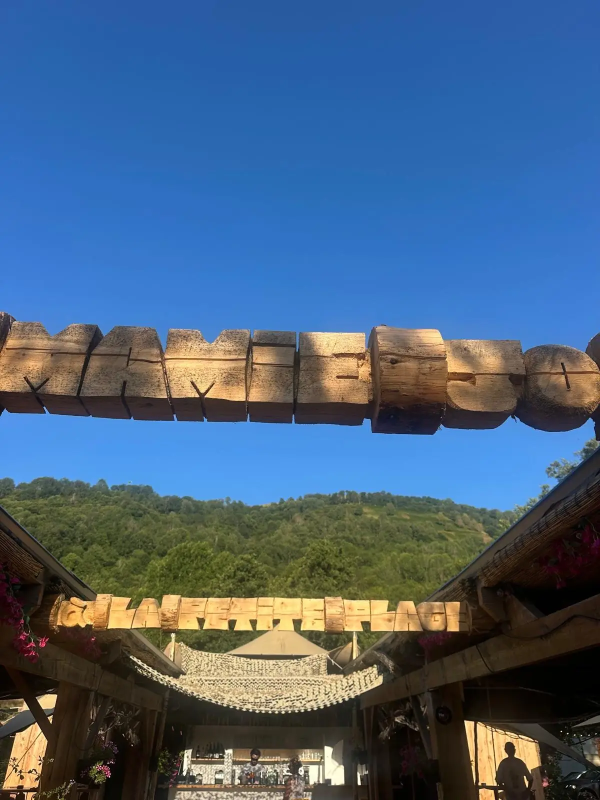 Portique en rondins de bois gravé « Mamie Jo » à l'entrée, sous un ciel bleu, montagnes boisées au fond