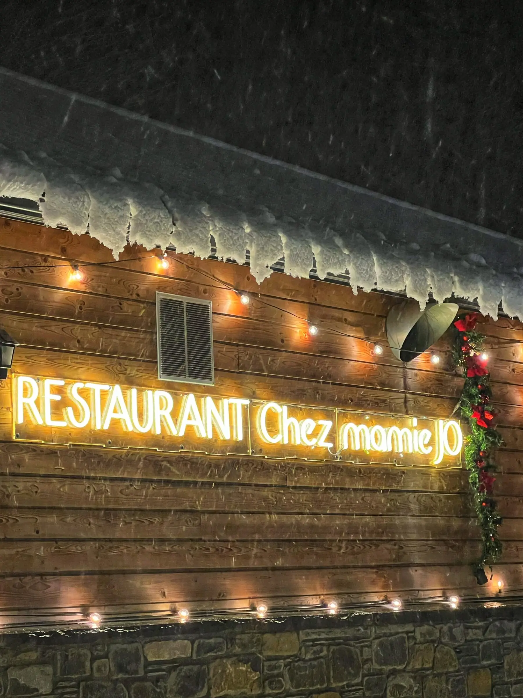 La façade en bois du chalet sous la neige, l'enseigne lumineuse « Restaurant Chez Mamie Jo » allumée