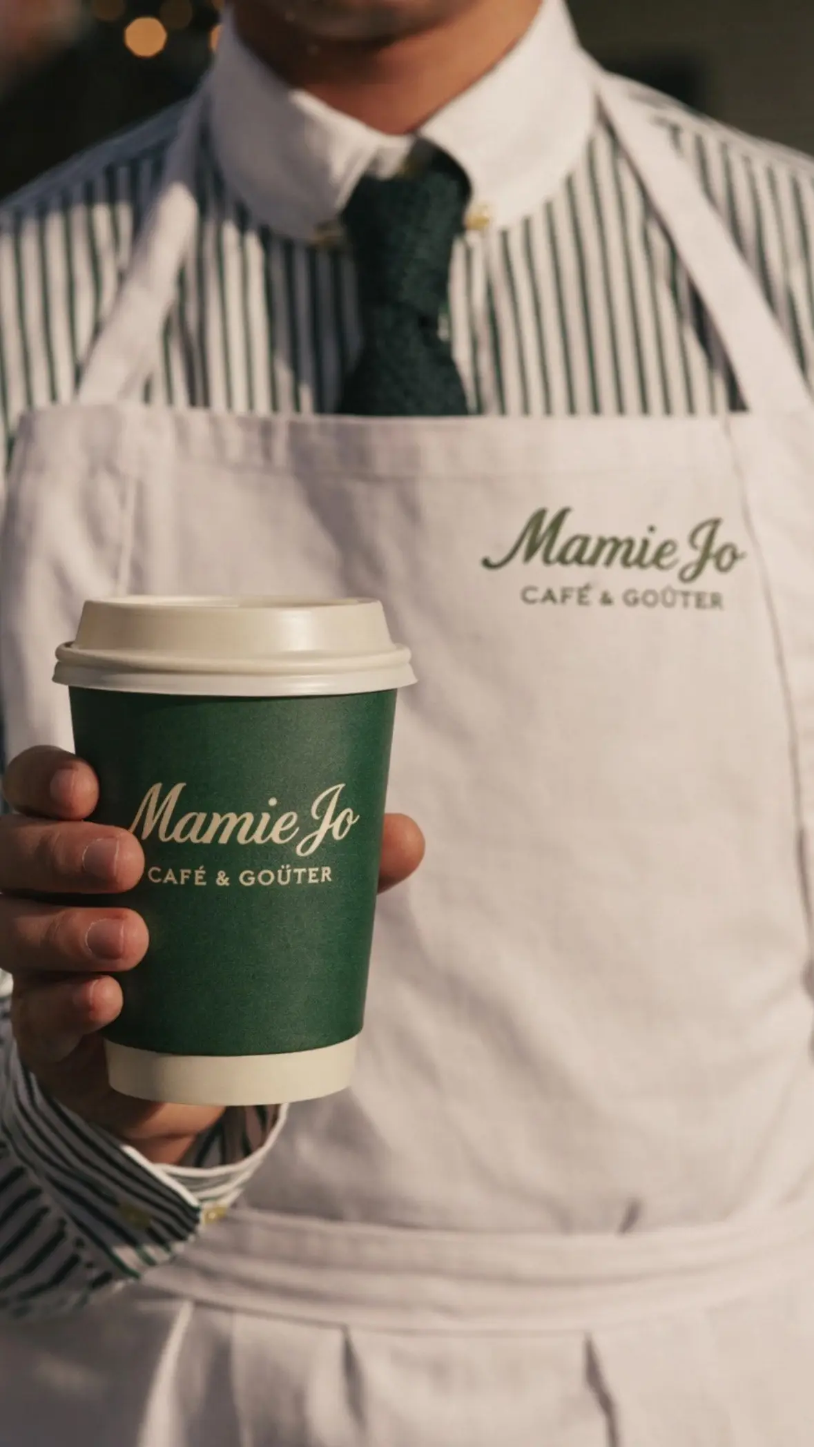 Gobelet et tablier aux couleurs de Mamie Jo Café & Goûter, tenus par un serveur