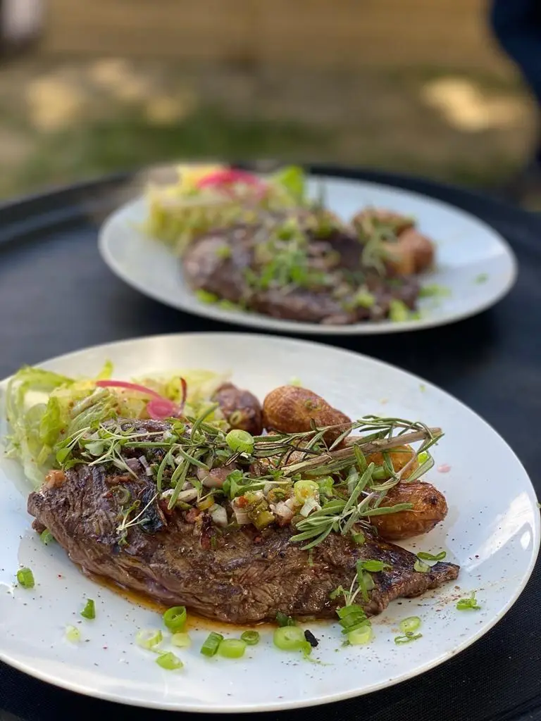 Pièce de bœuf grillée, pommes grenaille, salade et pickles d'oignon rouge, servie en terrasse