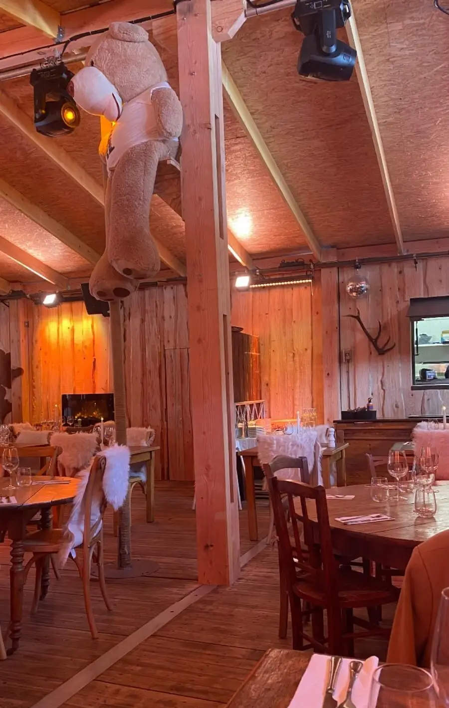 La salle du chalet tout en bois, tables dressées, chaises couvertes de peaux et grand ours en peluche accroché à une poutre