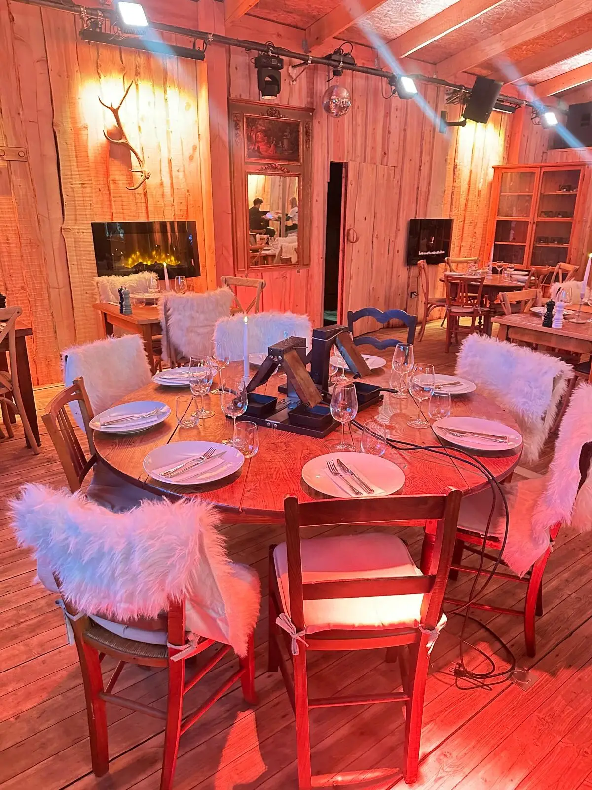 Grande table ronde dressée sous une lumière rouge, chaises couvertes de fourrure blanche, trophée de cerf au mur