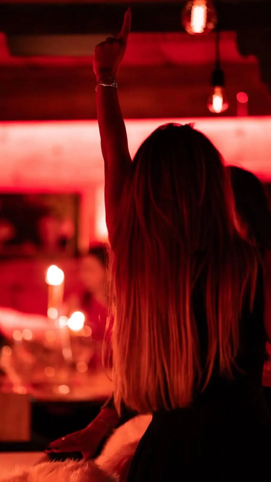 Une invitée lève le bras en dansant, de dos, dans la salle baignée de lumière rouge, bougies allumées sur les tables